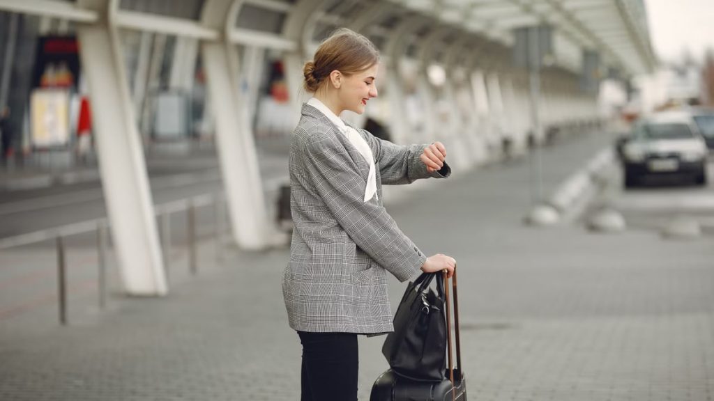 Side view of happy woman in checkered jacket standing with luggage and looking on watch before flight departure in airport